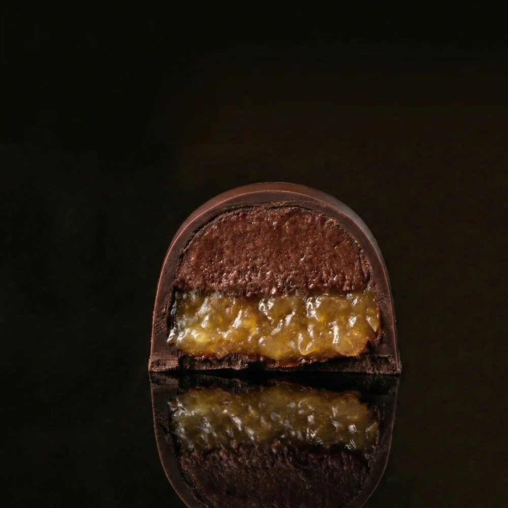Un bombón brillante cortado por la mitad que revela un gel translúcido de maracuyá sobre una ganache oscura.